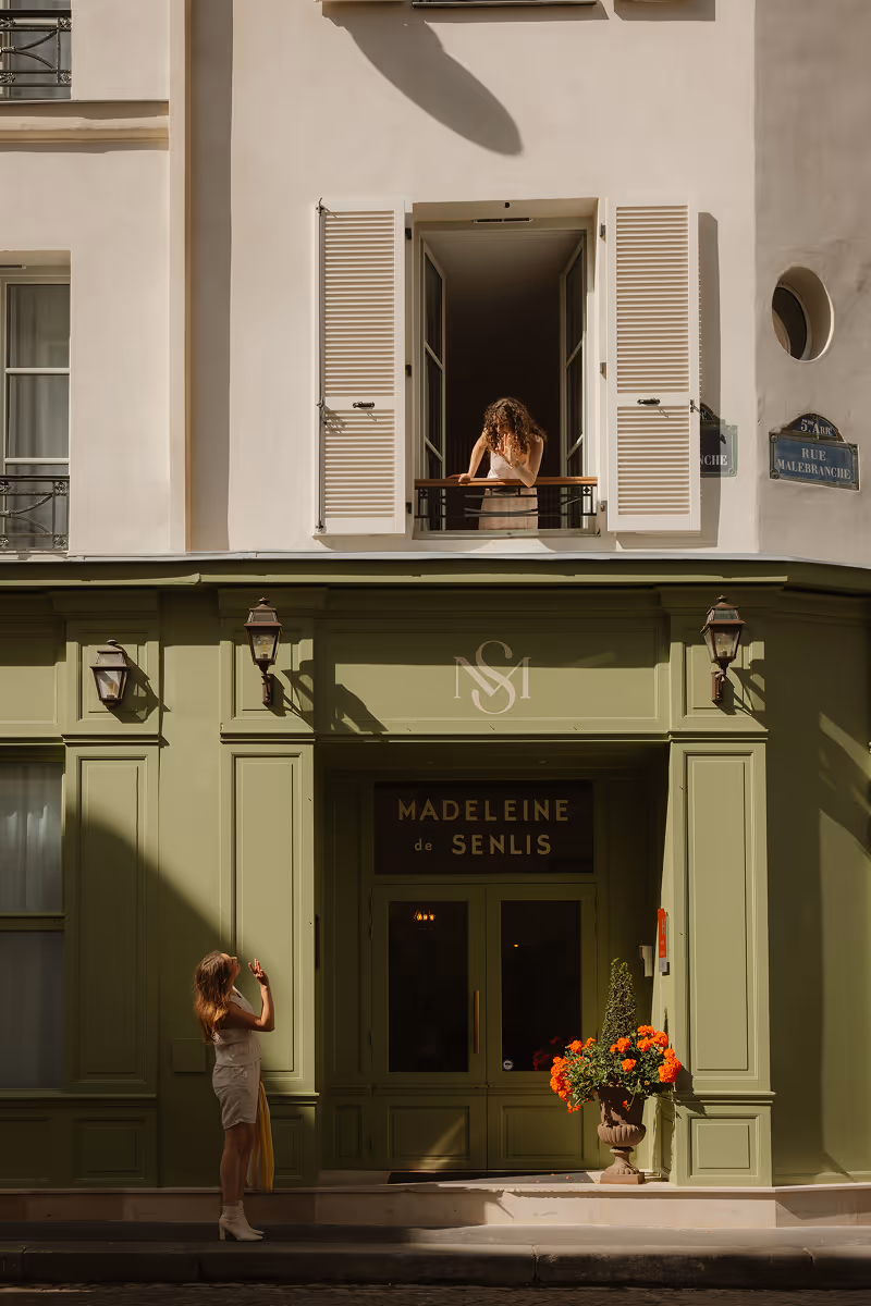 Une femme parle à une autre au balcon au-dessus d'une boutique verte nommée Madeleine de Senlis.