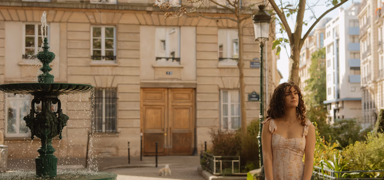 Une femme aux cheveux bouclés en robe claire se tient près d'une fontaine verte dans une rue urbaine.