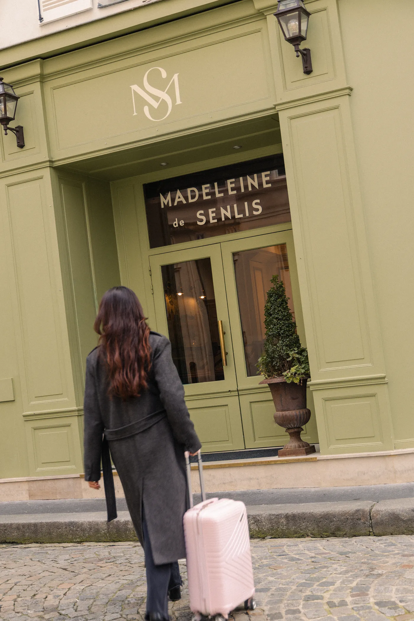 Femme vue de dos avec valise rose marchant vers l'entrée verte de Madeleine de Senlis.