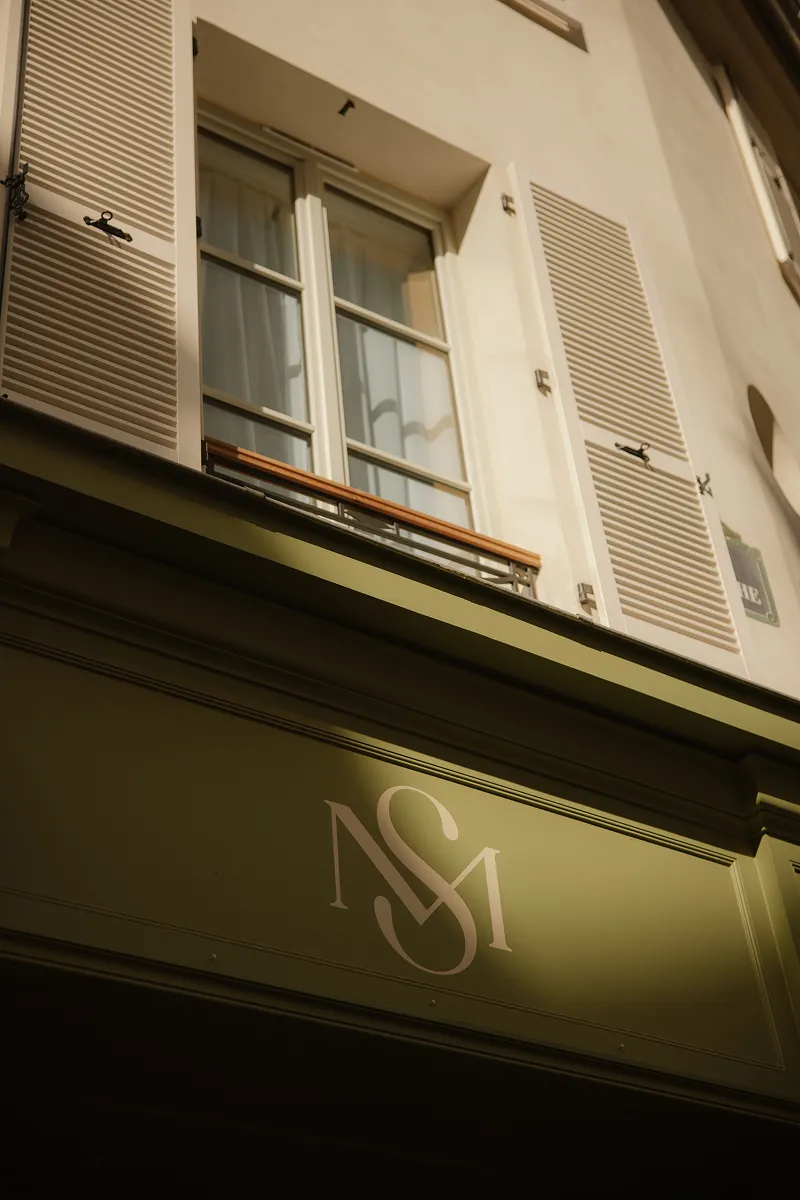 Façade créme avec fenêtres à volets blancs ouverts et un panneau vert olive avec logo NSM.