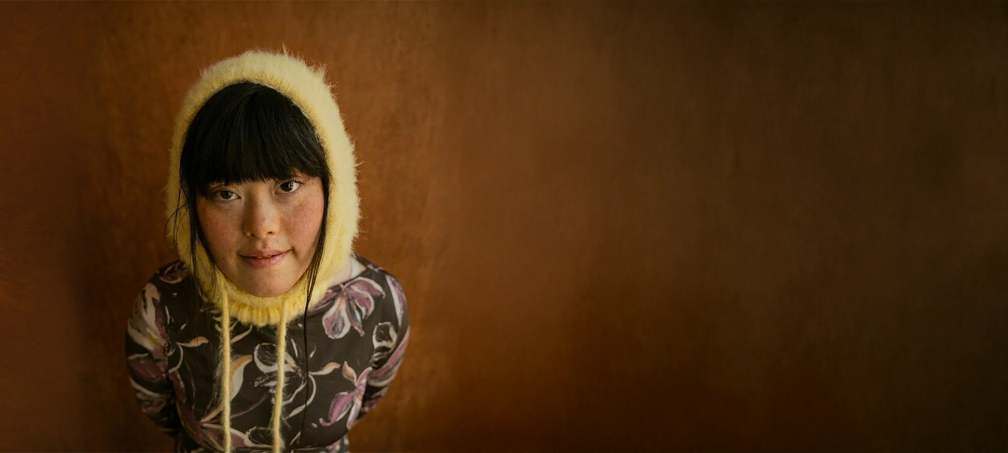 Young woman with black hair wearing a yellow knit hood and floral patterned shirt looking at the camera.