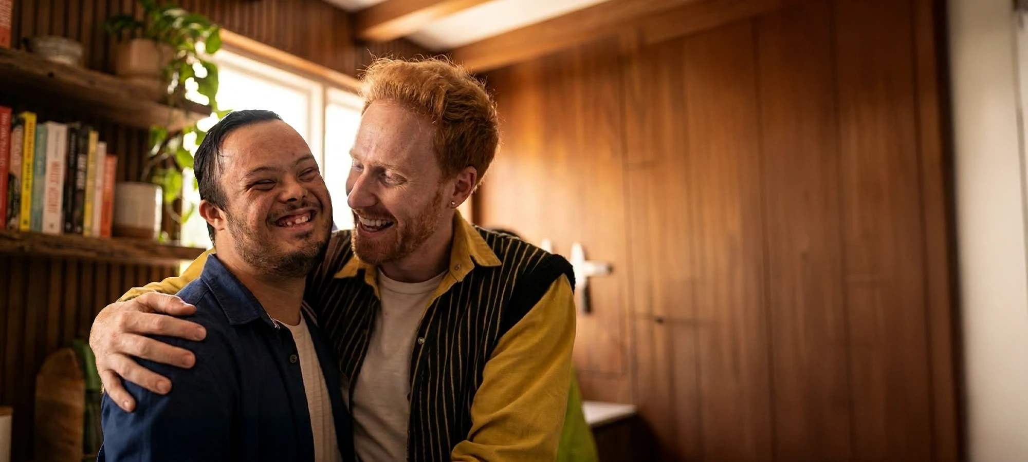 Two men warmly embracing and smiling in a cozy, wood-paneled room with a bookshelf and plant.