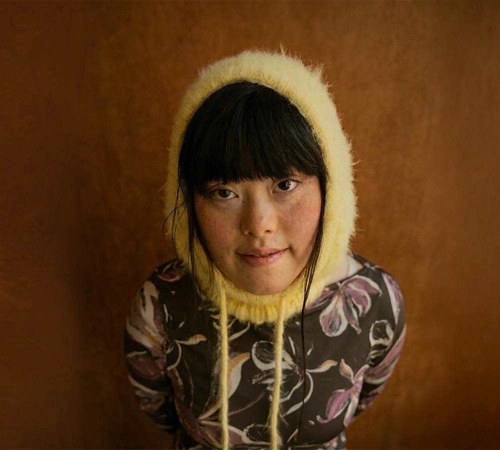 Woman with dark hair wearing a yellow knitted bonnet and floral-patterned top against a brown background.