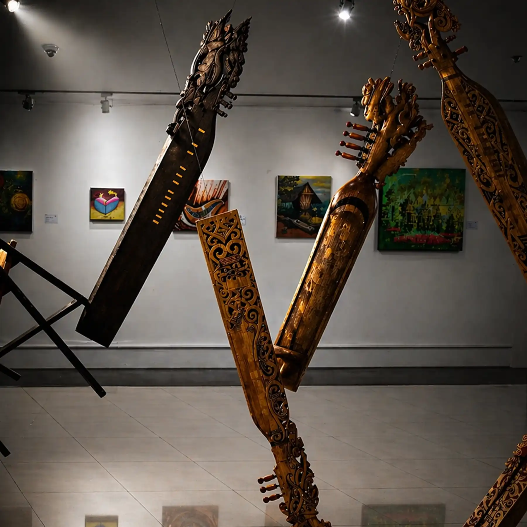 Pameran galeri dengan alat musik tradisional ukiran kayu besar tergantung dan lukisan warna-warni di dinding.