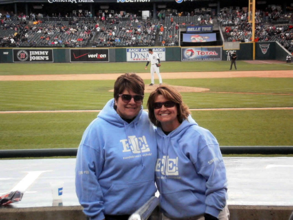 Two women in blue hoodies and sunglasses smiling at a baseball stadium with a player on the field.