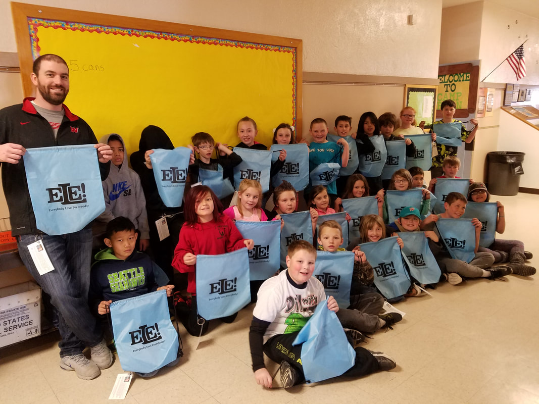 Group of children and an adult holding blue bags that say 'Everybody Love Everybody!' in a school hallway.