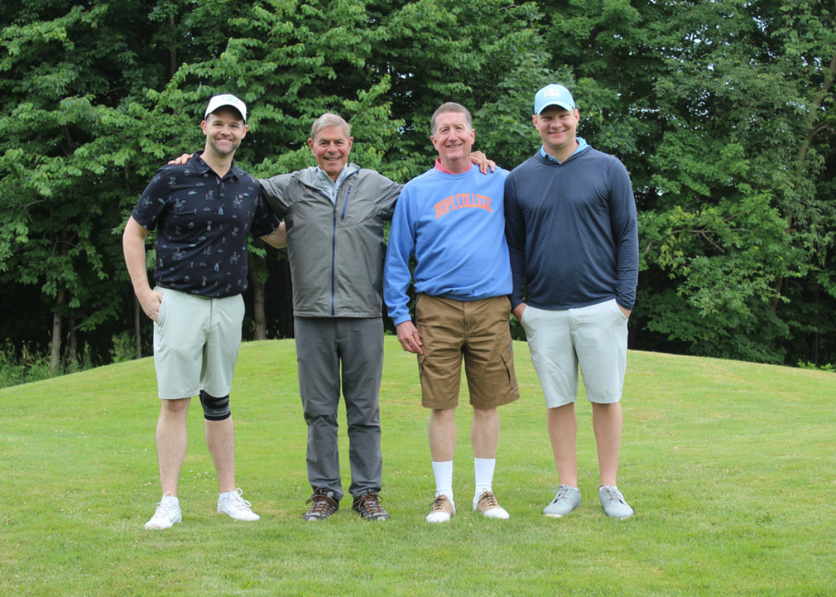 Four men standing on grass with trees behind, three wearing caps and casual summer clothing.