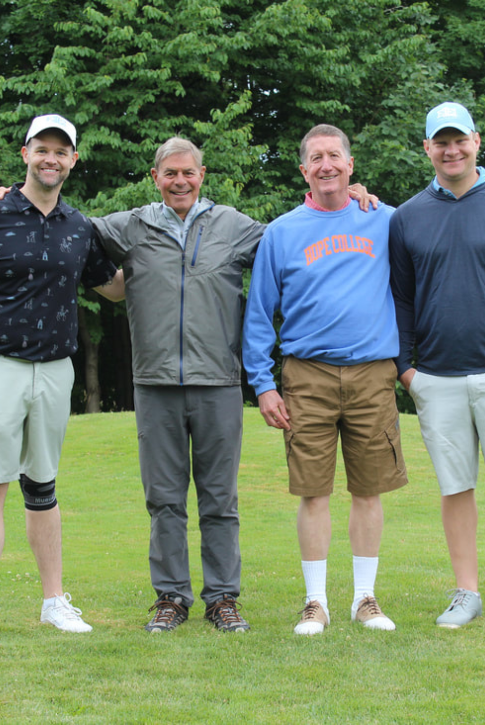 Four men standing arm in arm on golf course grass with trees in background, smiling at camera.