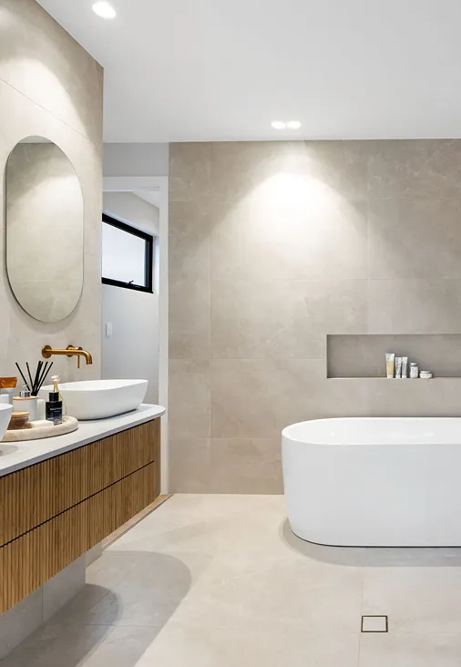 Modern bathroom in a PlanCo Homes custom build, featuring a freestanding white bathtub, beige floor and wall tiling, a timber vanity with vessel basin and oval mirror