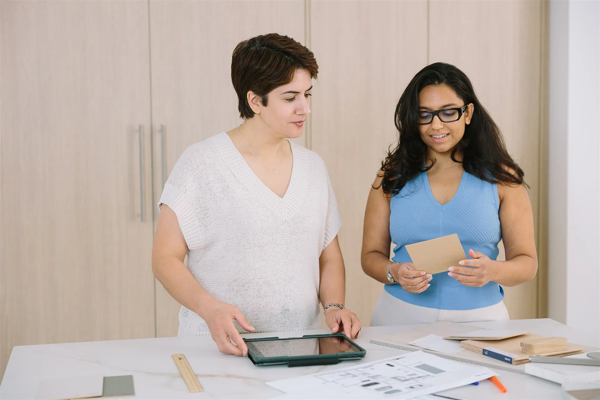 Two women reviewing materials and documents on a table, one holding a tablet and the other examining a square sample.