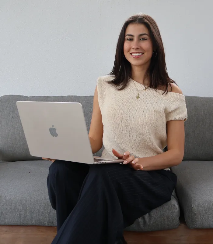 Smiling woman with dark hair sitting on a gray couch using a silver Apple laptop, wearing a beige off-shoulder top and dark pants.
