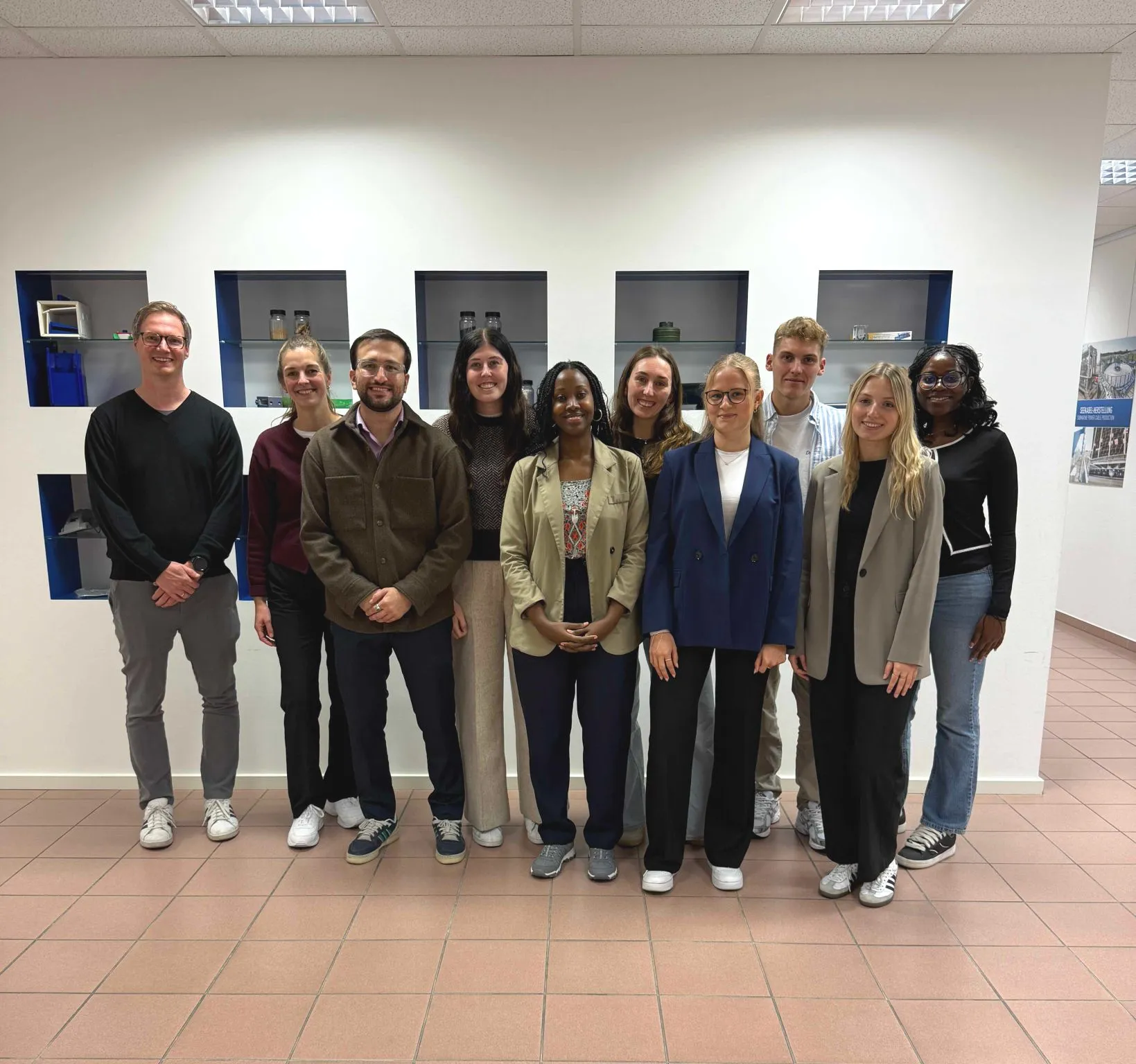 Group of ten diverse young adults standing indoors against a white wall with display niches.