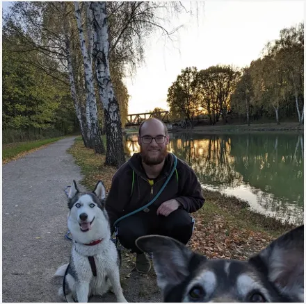 Man crouching on path by a river with a husky dog sitting beside him and another dog in foreground.
