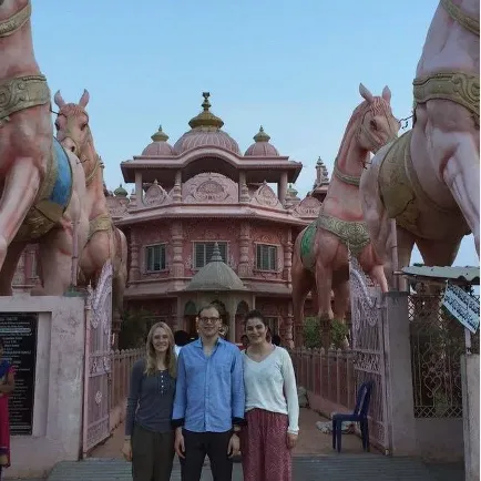 Three people stand in front of a pink temple entrance flanked by large horse statues under a clear sky.