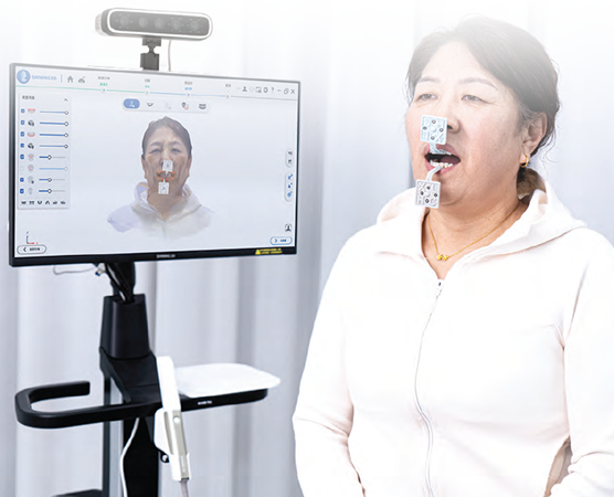 Woman wearing markers on face standing near a monitor displaying a 3D facial scan.