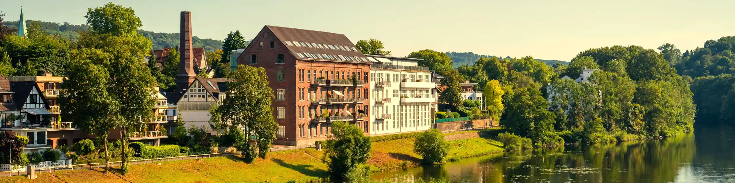 Blick auf ein saniertes Fabrikgebäude mit modernen Balkonen am ruhigen Flussufer in Essen, umgeben von viel Grün und angrenzender Wohnbebauung.