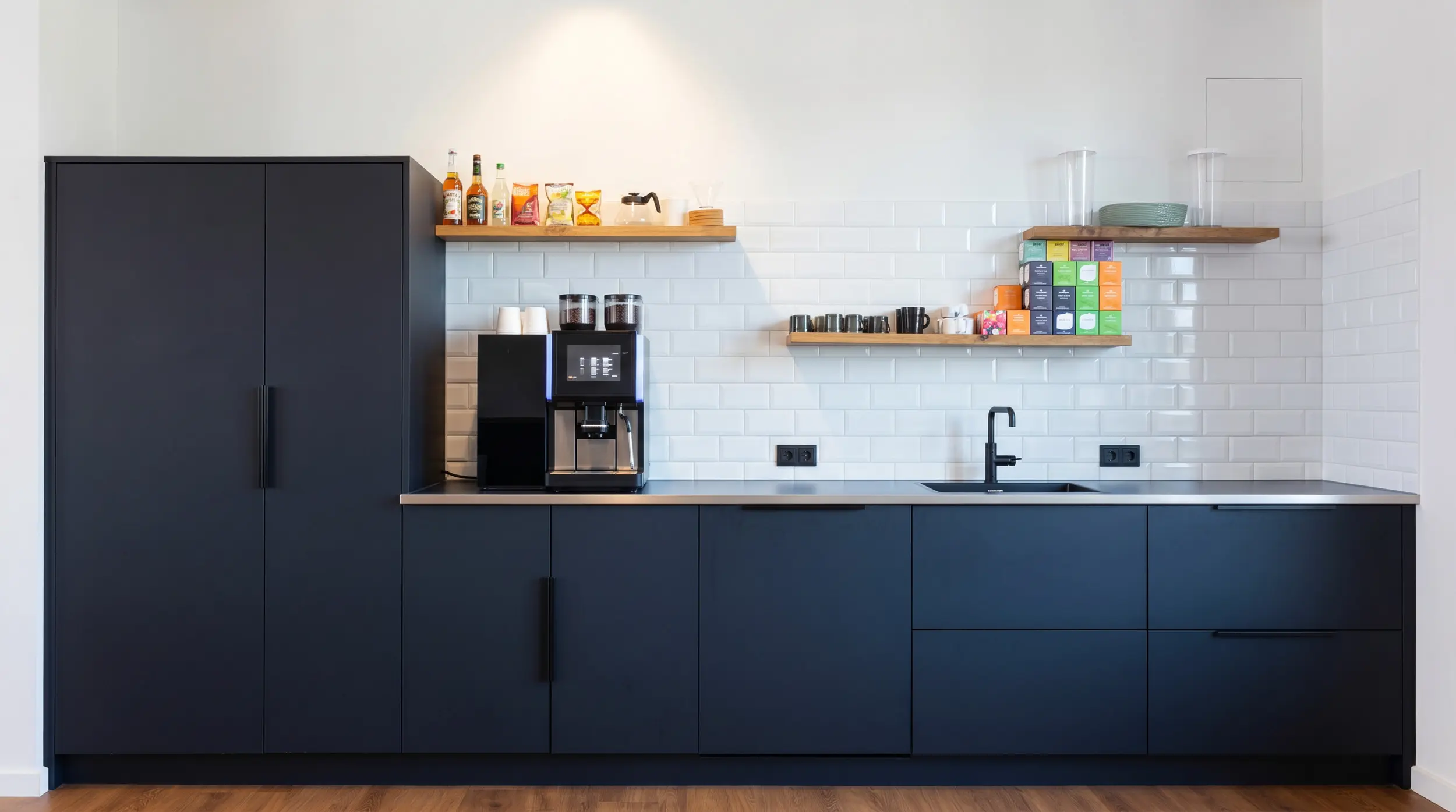 Moderne Office-Kaffeeküche mit dunklen Fronten, Spüle und Kaffeemaschine vor Fliesenwand.