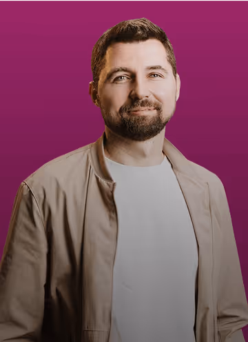 Mann mit Bart, hellem T-Shirt und beiger Jacke vor violettem Hintergrund, lächelnd.
