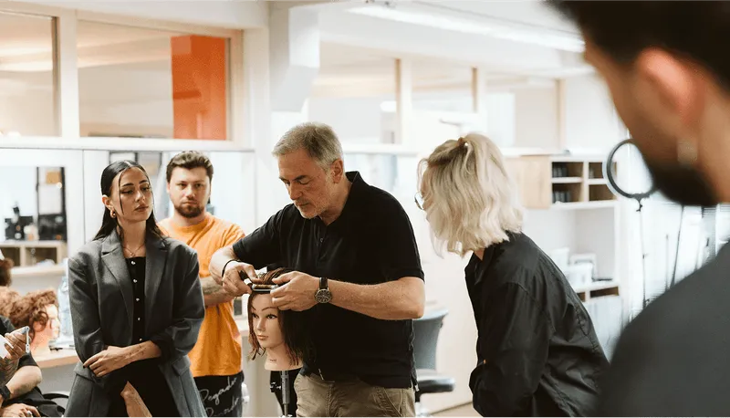 Frisörkurs: Mann schneidet Haarpuppe, andere Teilnehmer beobachten konzentriert im Salon.