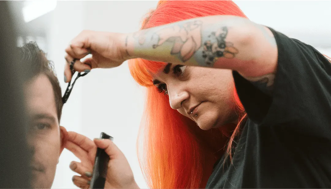 Friseurin mit rotem Haar und Tattoos schneidet konzentriert die Haare eines Kunden in einem modernen Salon.