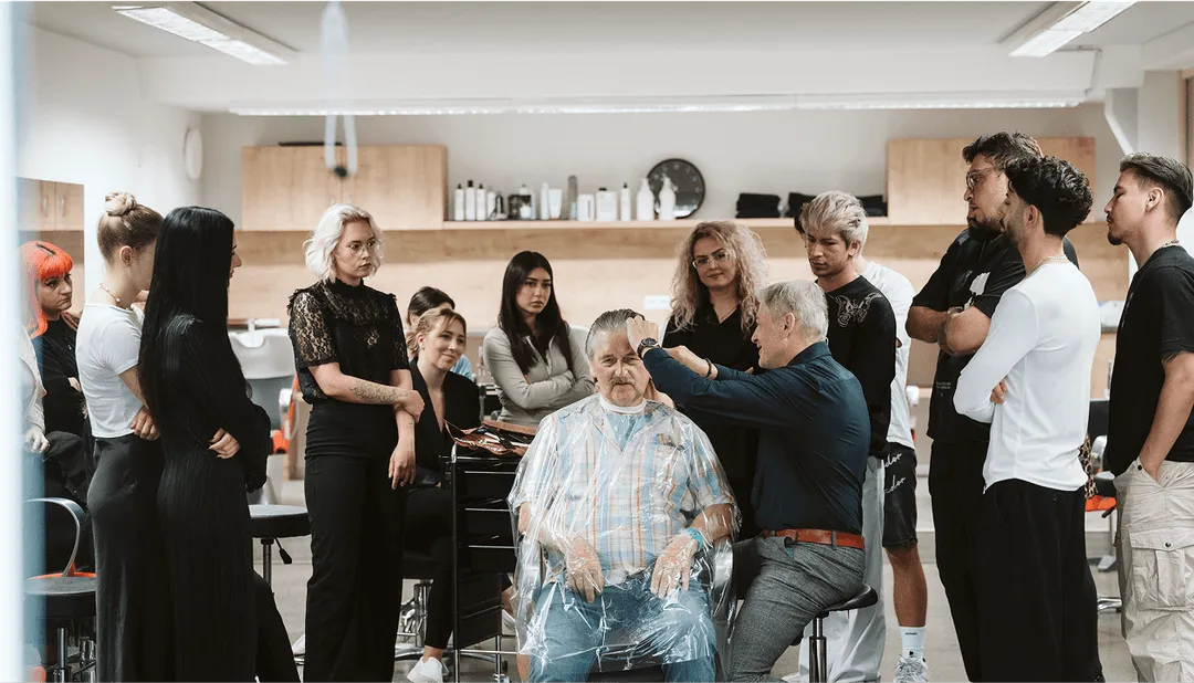 Friseur lehrt Haarschneidetechniken umringt von Lehrlingen, während älterer Mann auf Stuhl sitzt. Training in Friseursalon.