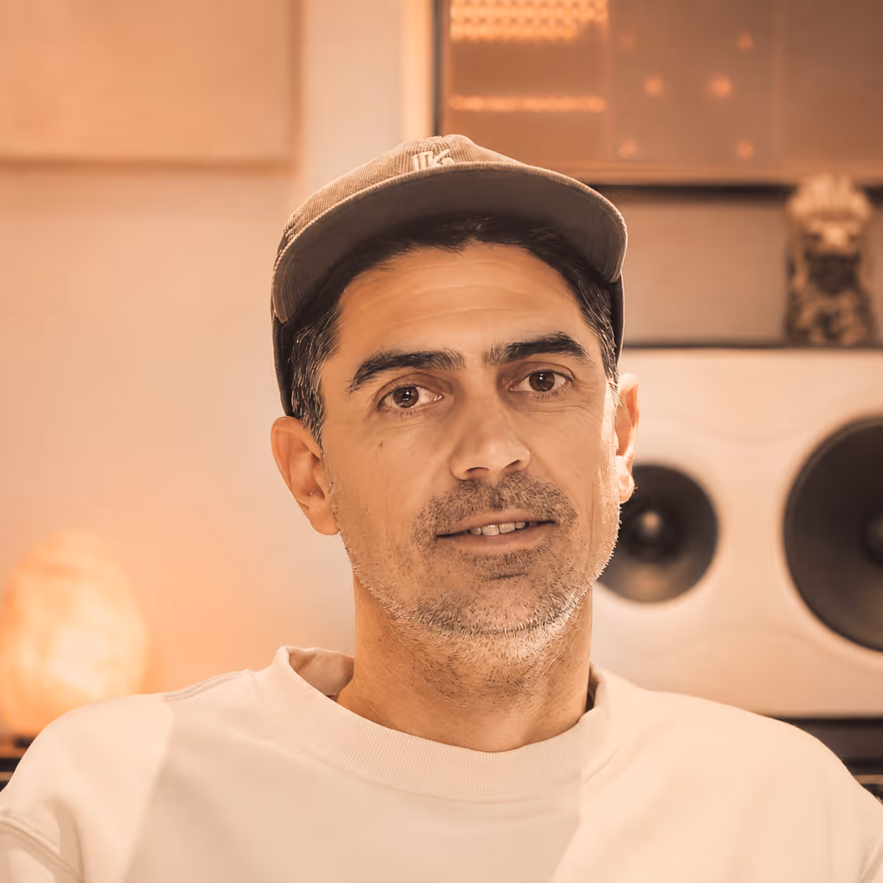 Man wearing a cap and light-colored sweatshirt in a warmly lit room with a large speaker in the background.