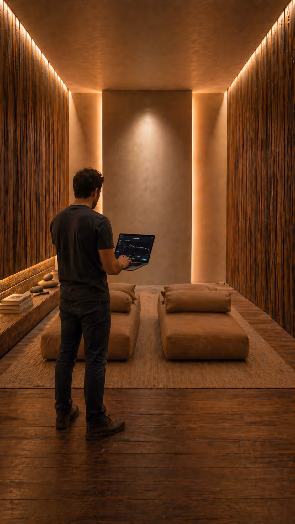Man in dark clothing standing in a warmly lit room with wooden walls, holding a laptop displaying a graph.