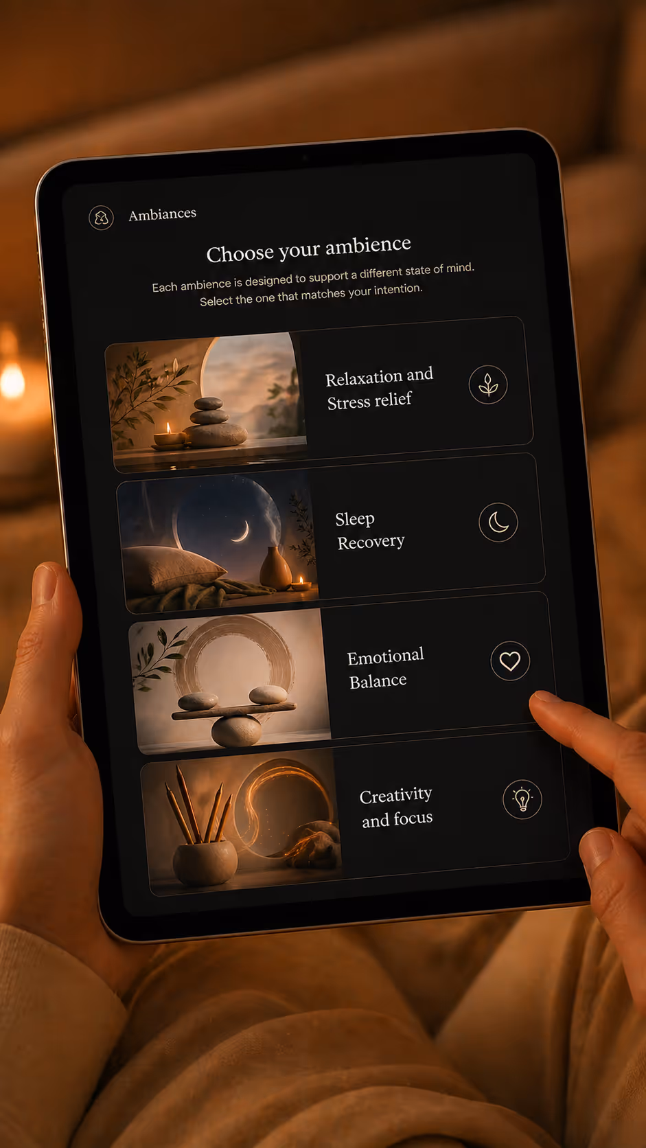Tablet screen displaying an ambience selection menu with options for Relaxation and Stress relief, Sleep Recovery, Emotional Balance, and Creativity and focus, held by a person touching the Emotional Balance option.