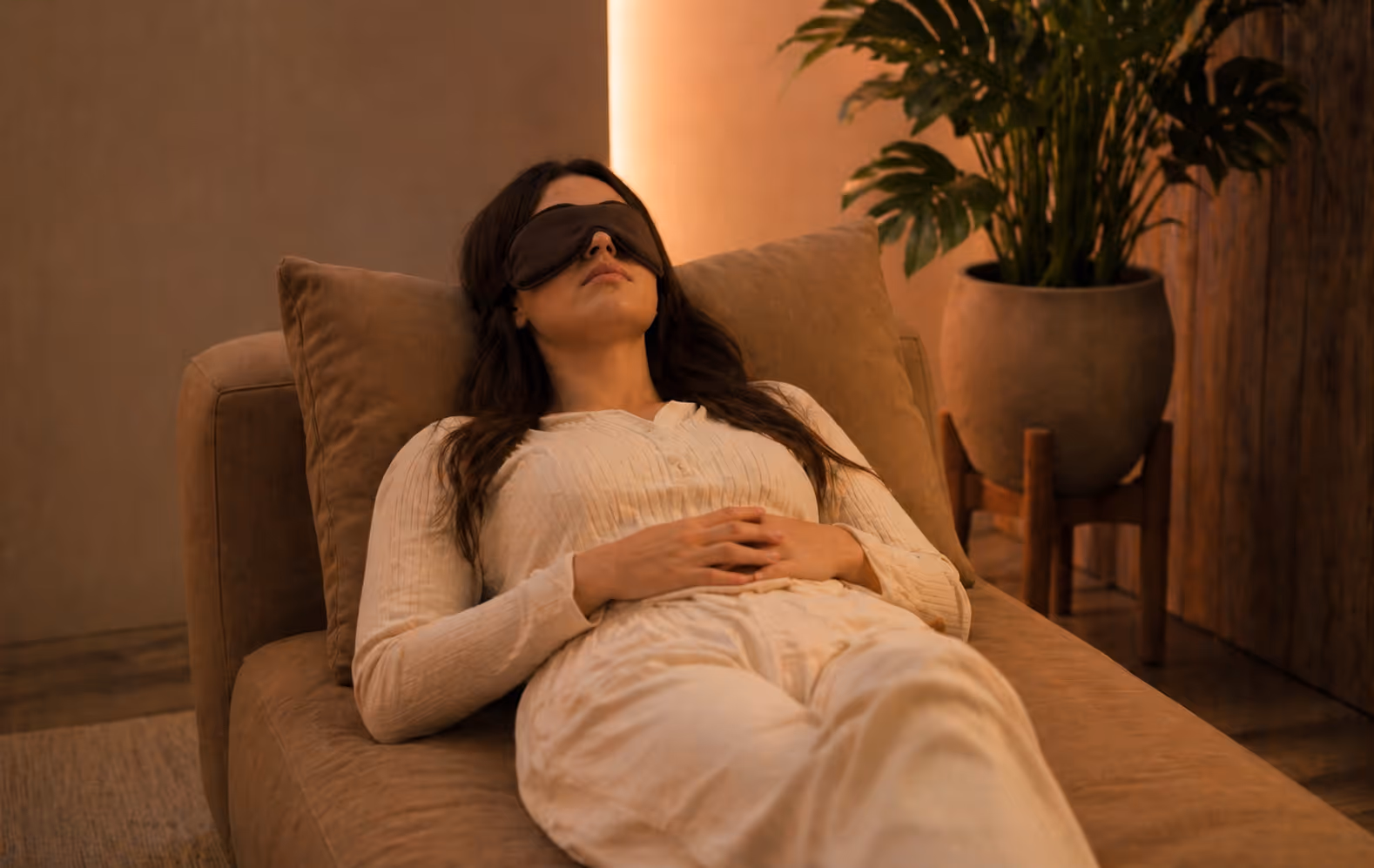 Woman reclining on a brown couch wearing a black sleep mask and white loungewear with hands resting on her stomach.
