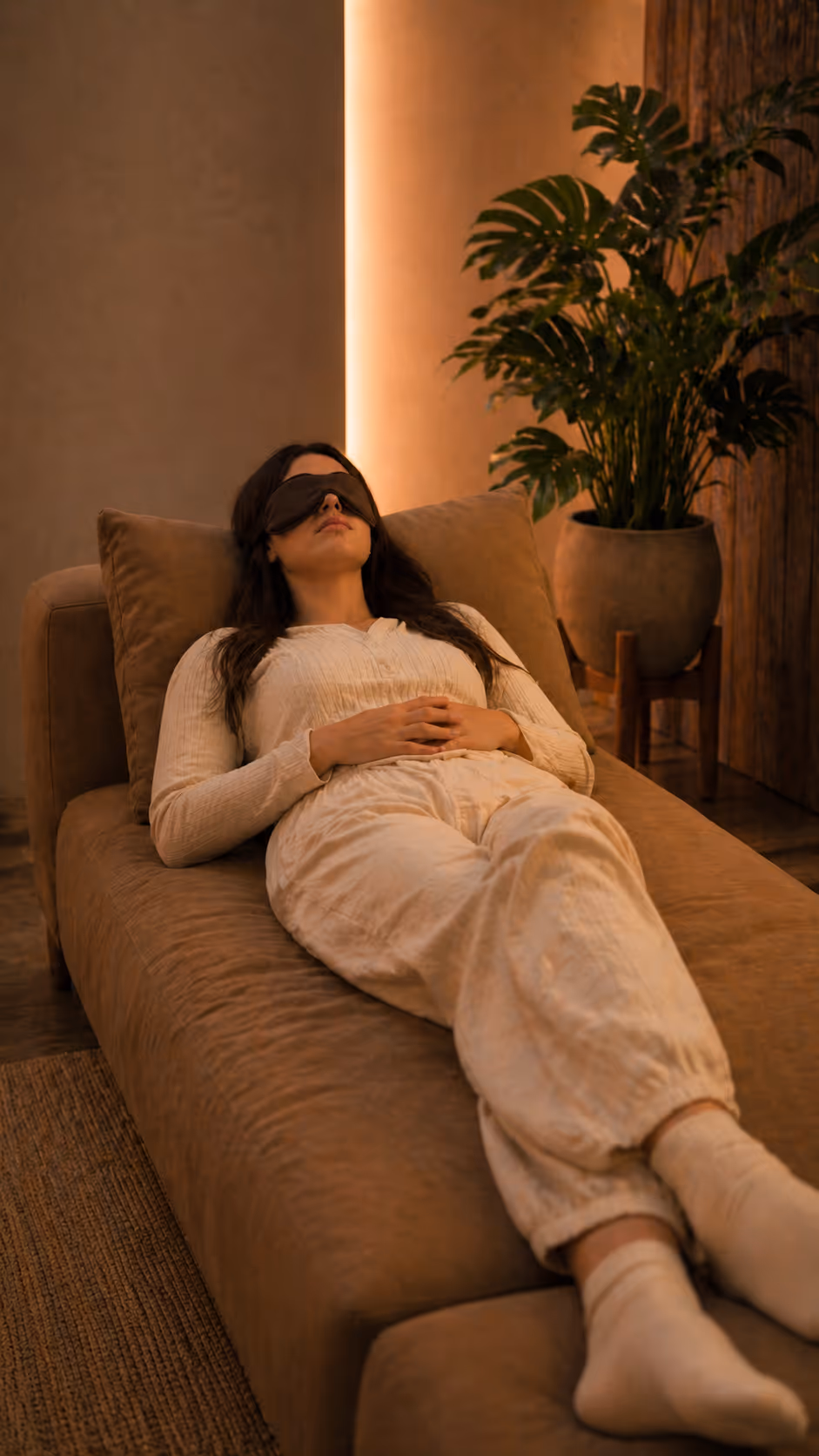Woman lying on a brown couch wearing an eye mask and beige comfortable clothing in a dimly lit room.