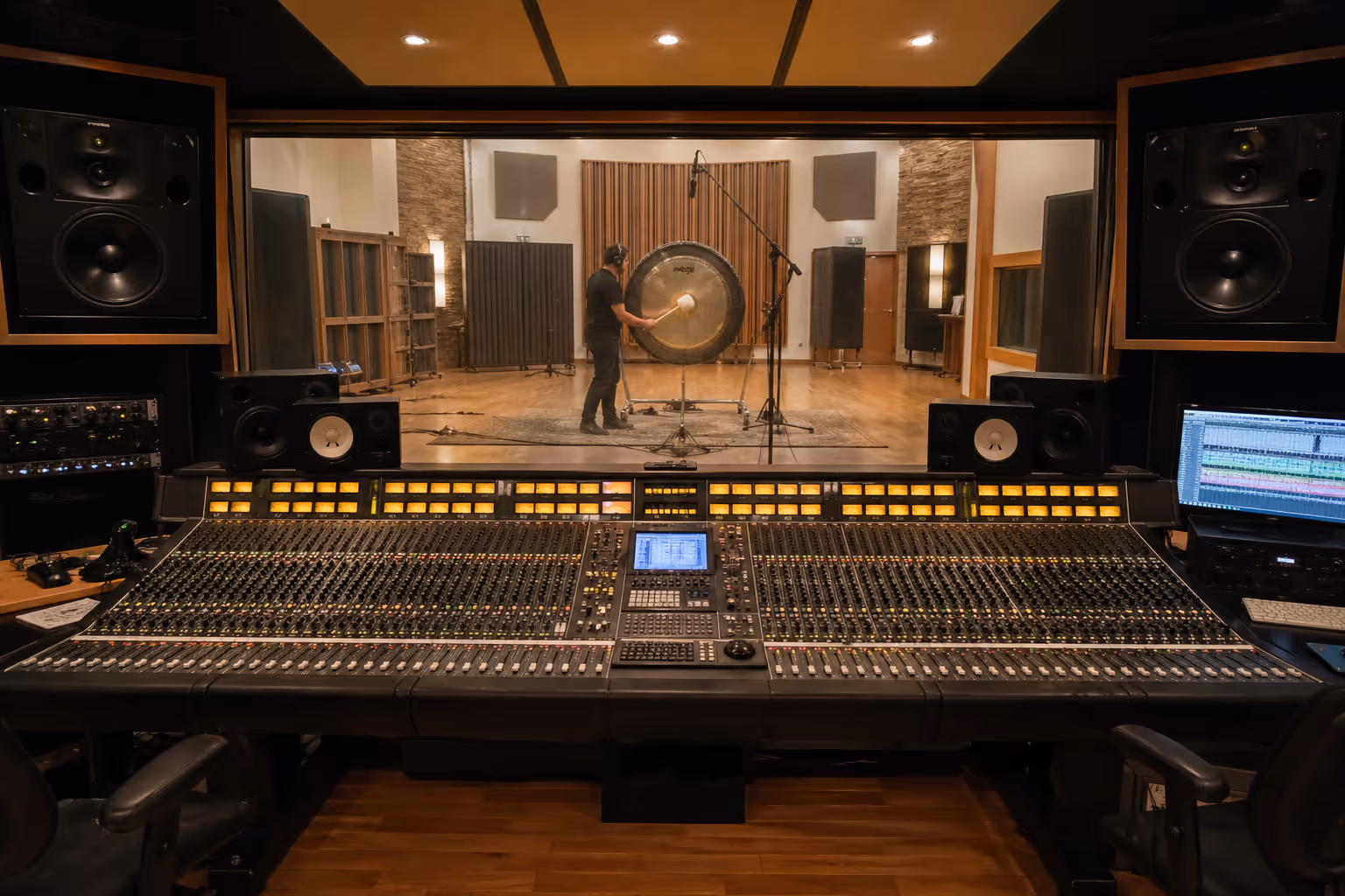 Audio mixing console in a recording studio with a man playing a large gong in the soundproof room visible through the glass window.