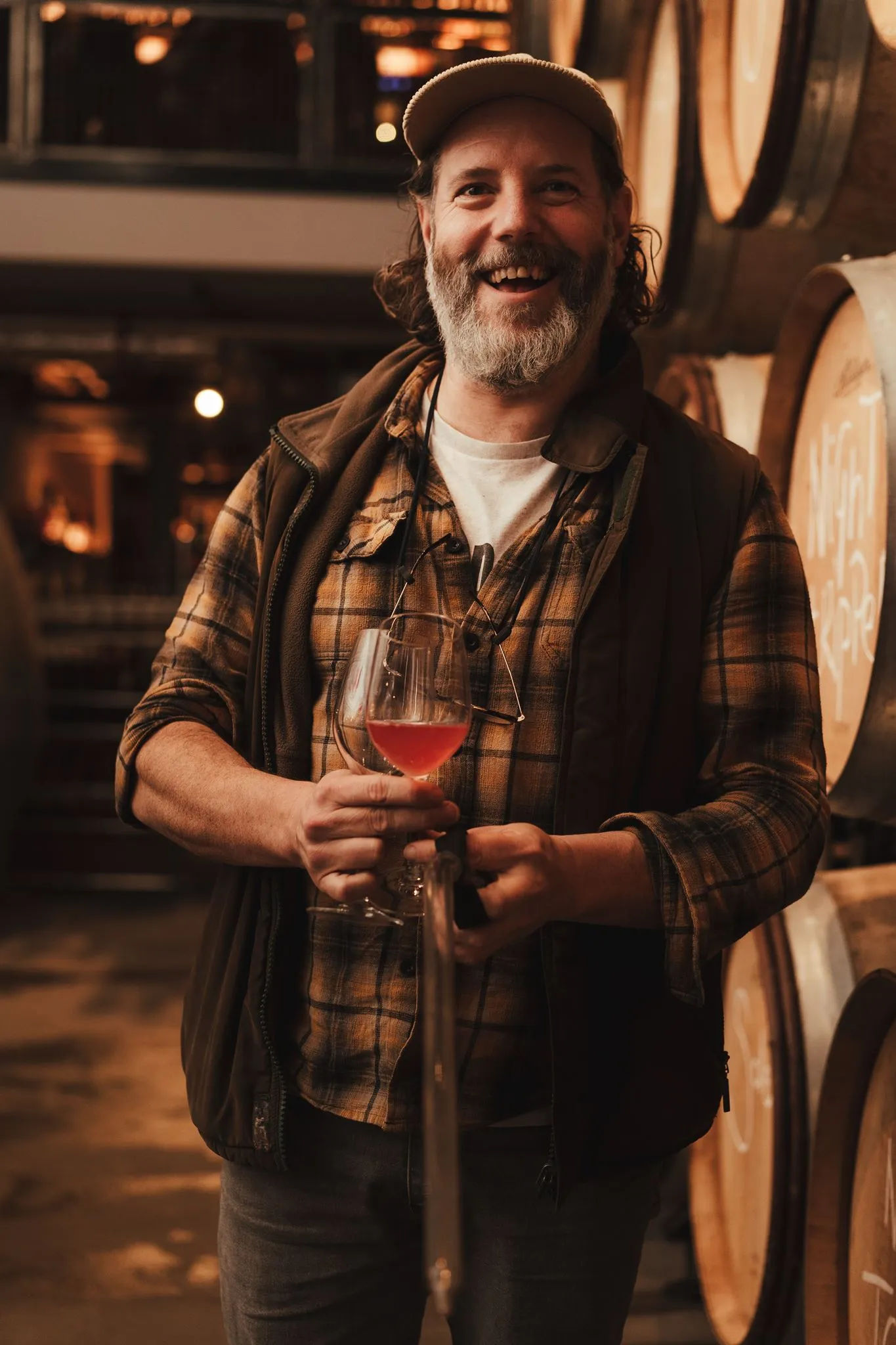 Smiling man with a glass of rosé among the oak barrels at the Vagabond Urban Winery