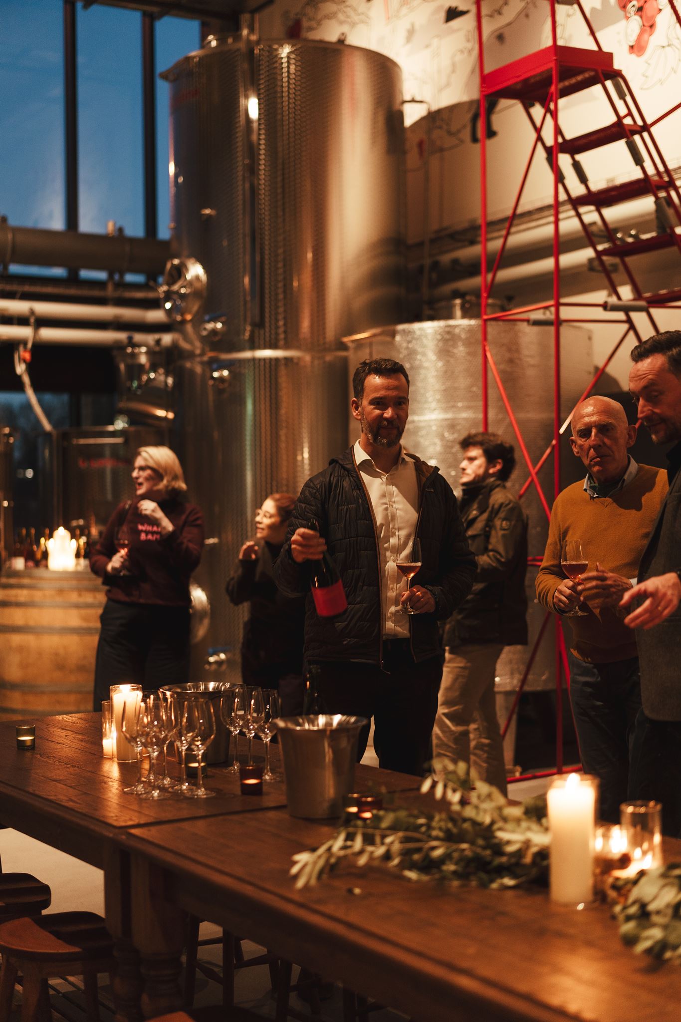 People socializing with wine glasses in a winery room with large metal fermentation tanks and candles.
