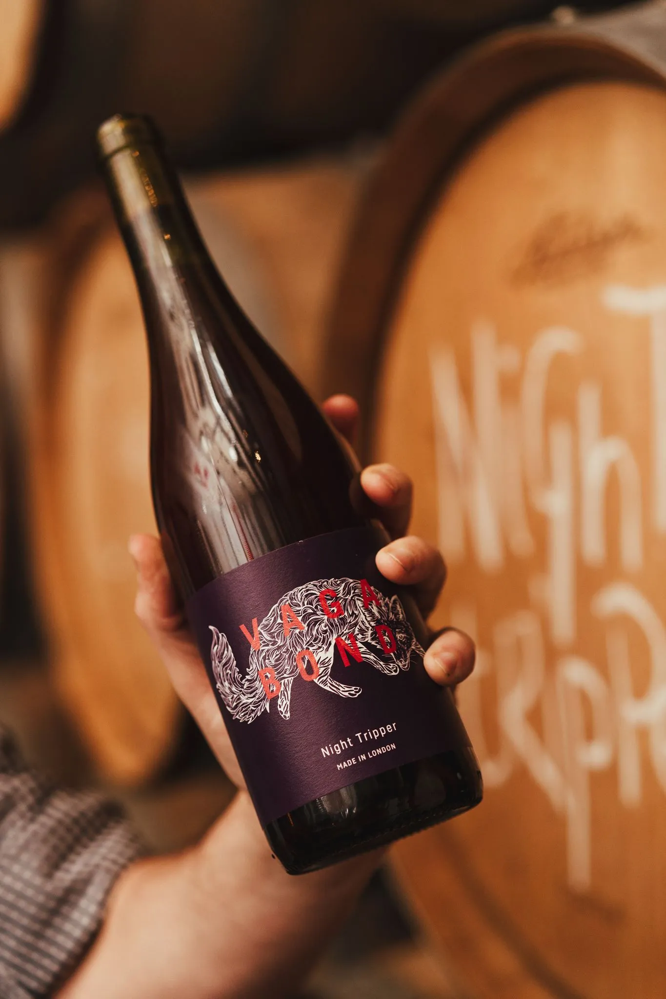 A bottle of Vagabond Winery Night Tripper held up in front of oak barrels