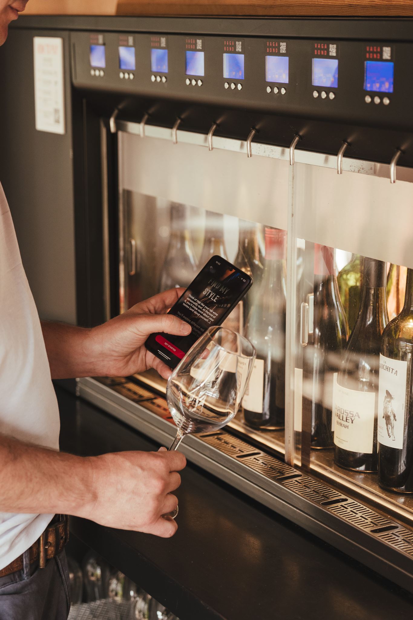 Person holding smartphone and wine glass near a self-serve wine dispenser with bottles inside.