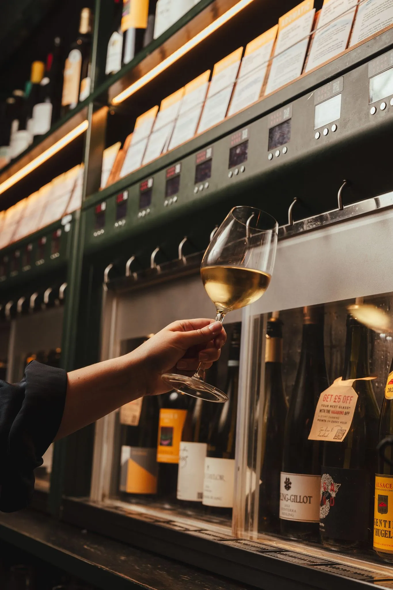 Glass of white wine held up beside a self-pour wine machine at Vagabond