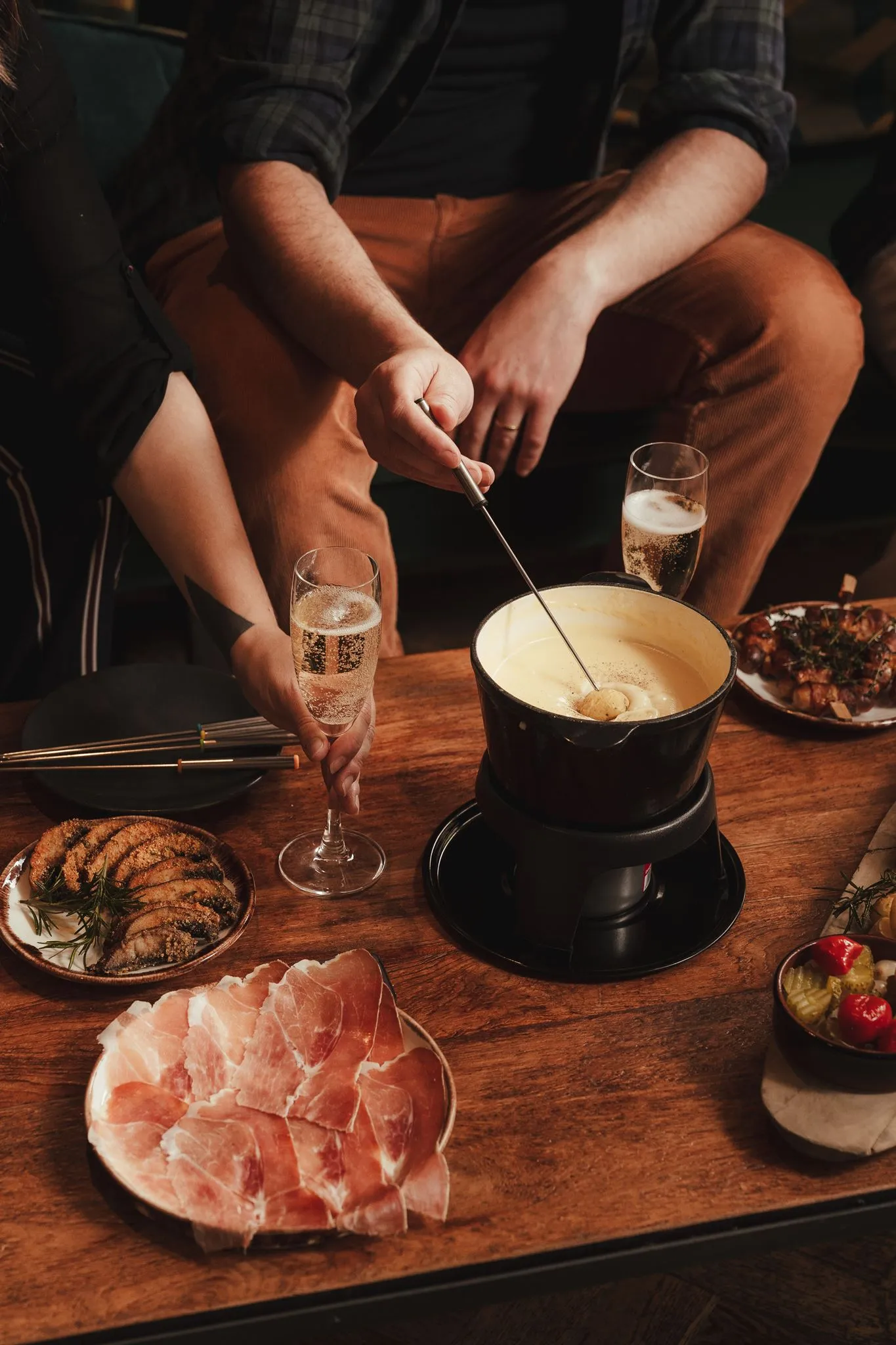 People dipping bread into cheese fondue with plates of cured meat, pickles, and sparkling drinks on wooden table.