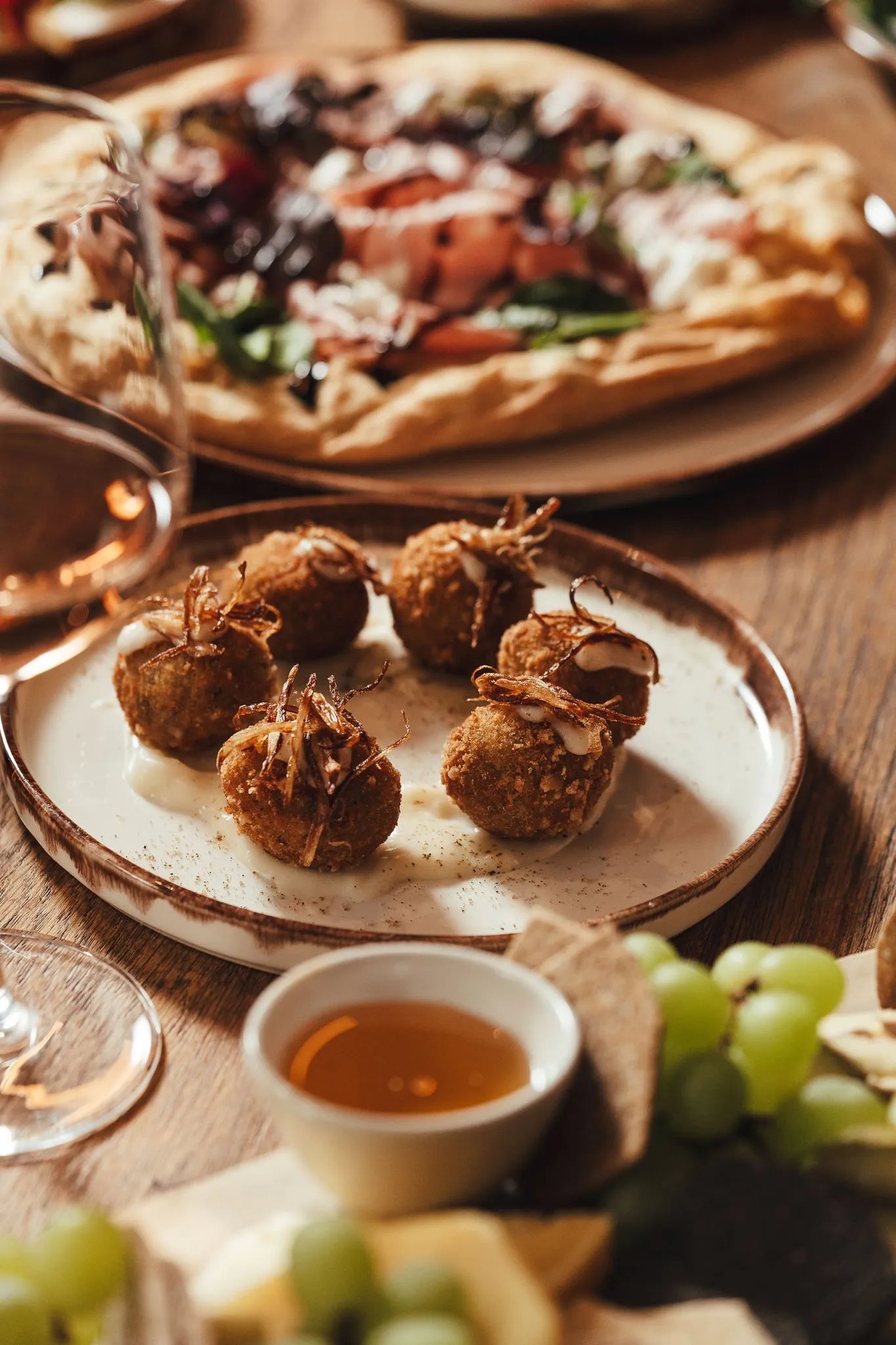 Plate with six breaded balls topped with crispy onions and sauce, with pizza and grapes blurred behind.