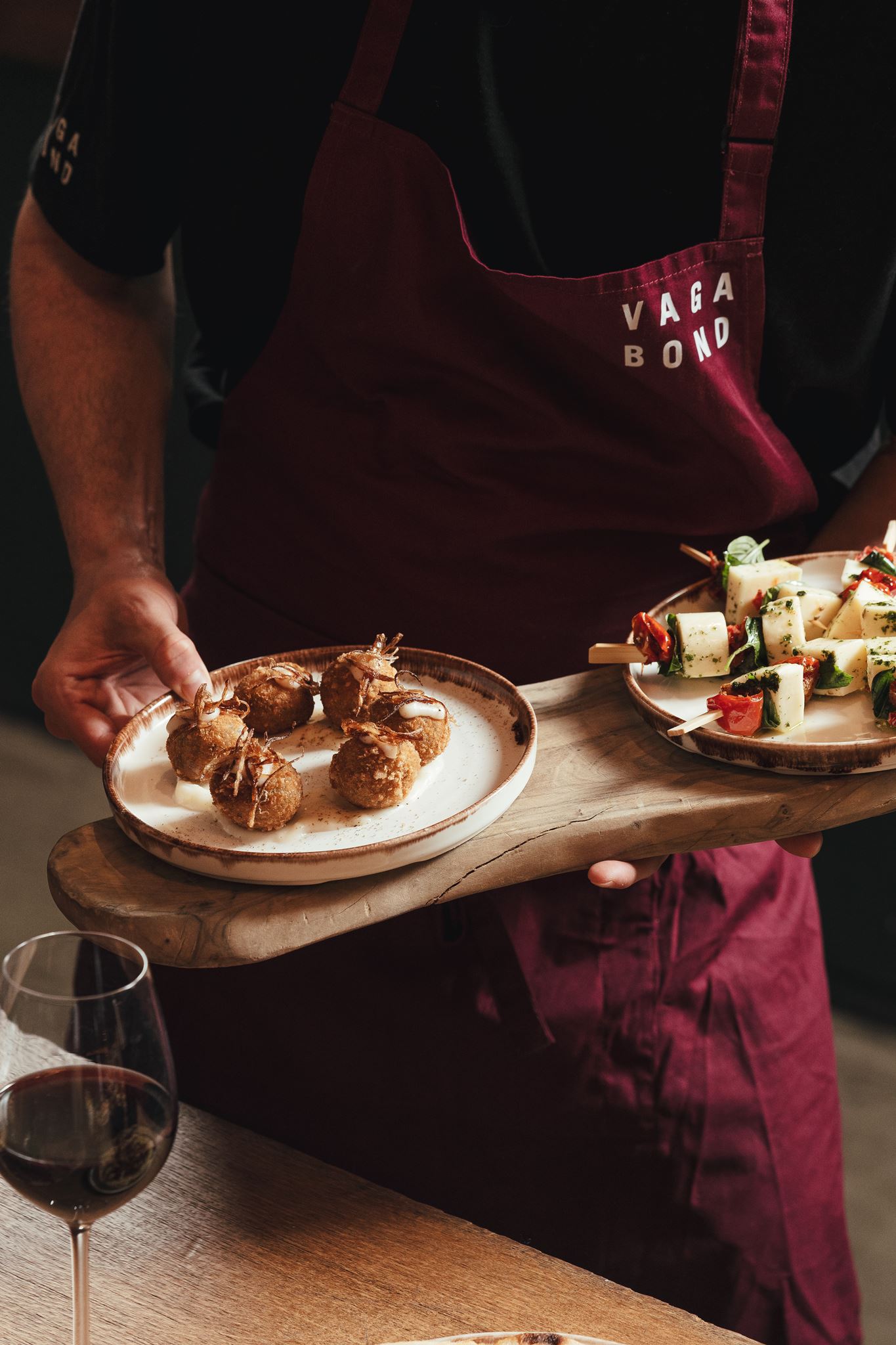 Person wearing a burgundy apron holds wooden board with two plates of appetizers and a glass of red wine nearby.