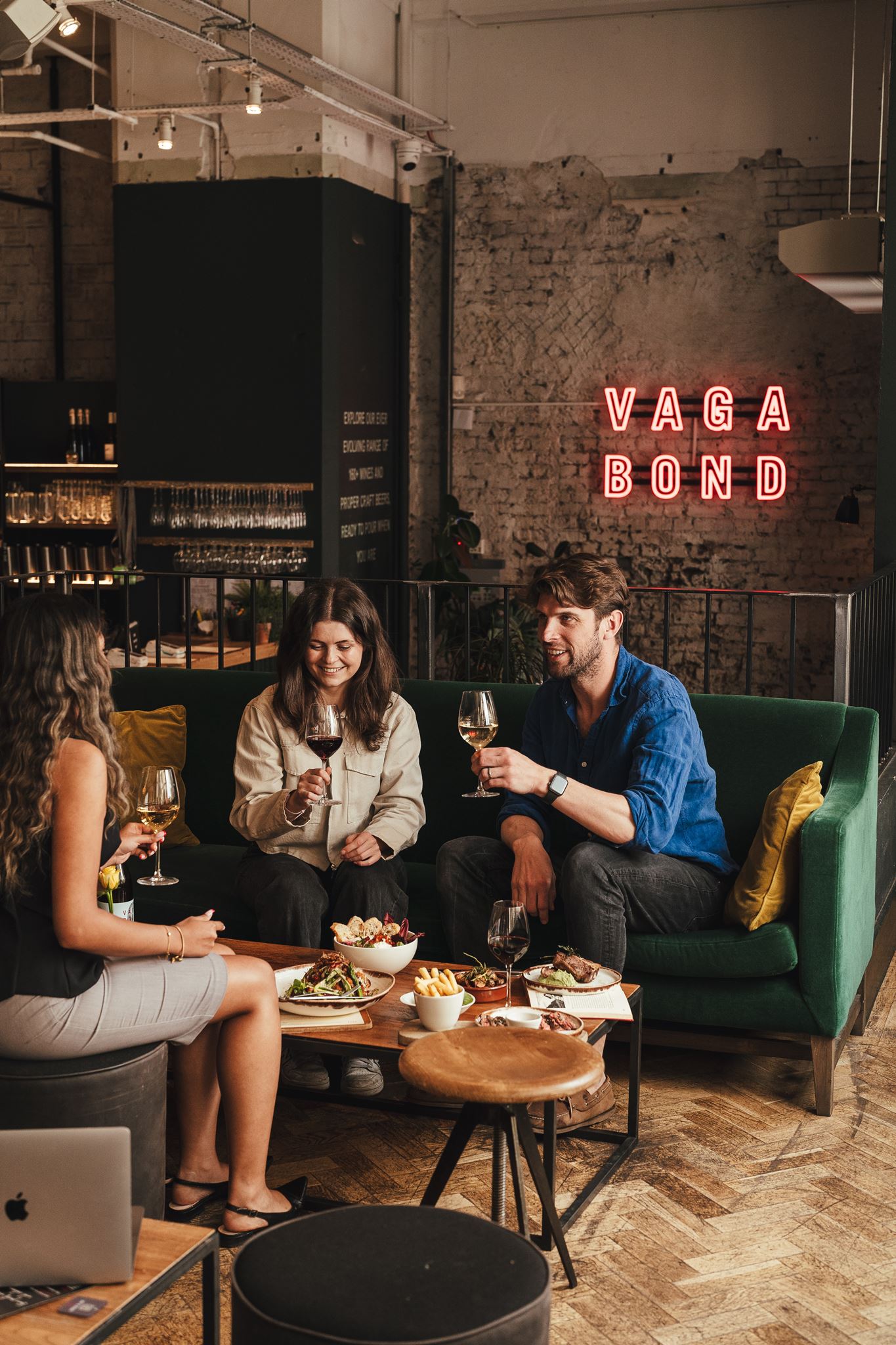 Three people sitting on green couch drinking wine and eating food in a cozy restaurant.