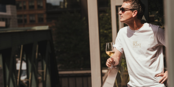 Man in sunglasses wearing a white t-shirt holding a glass of white wine outdoors in sunlight.