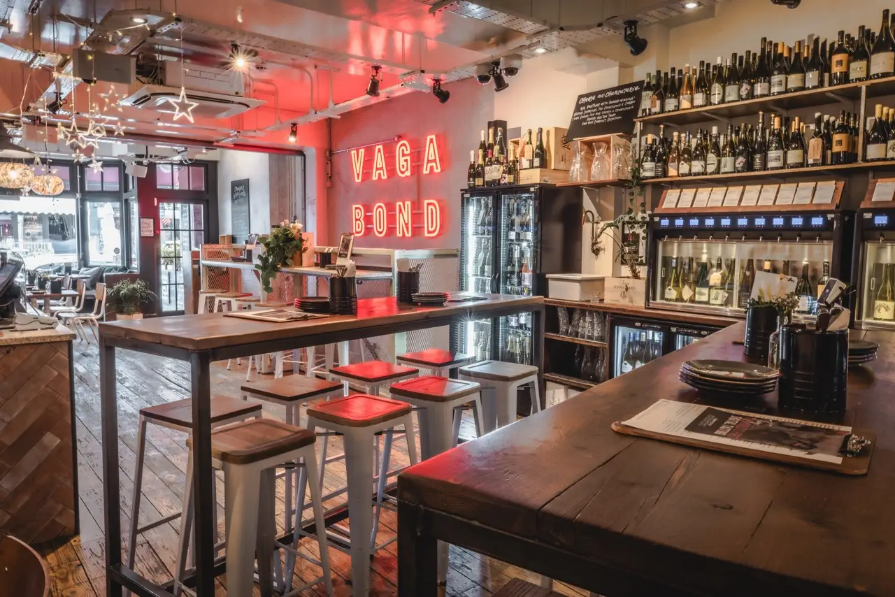 Red neon Vagabond sign above high tables and self-pour wine machines at Vagabond Charlotte Street