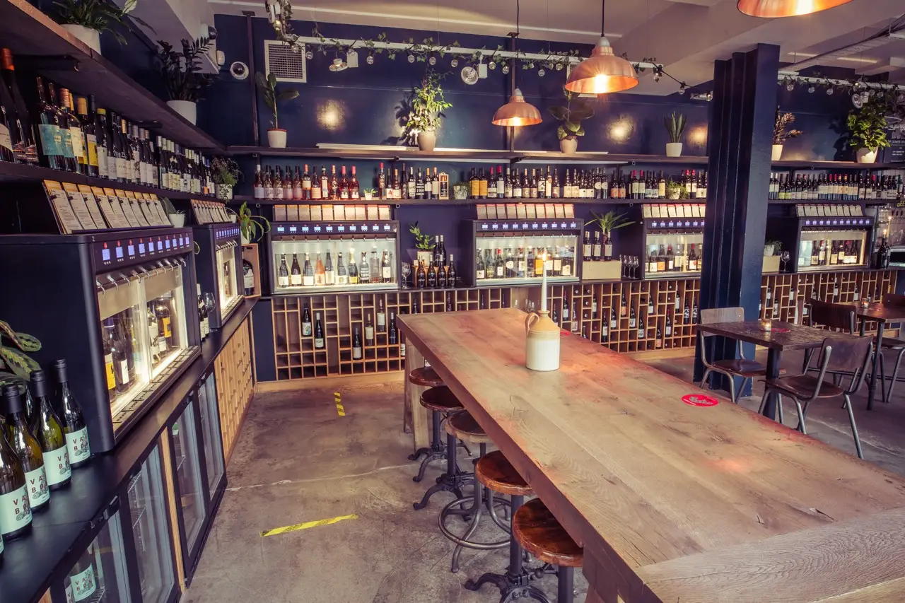 Long oak sharing table beside walls of self-pour wine machines at Vagabond Fulham