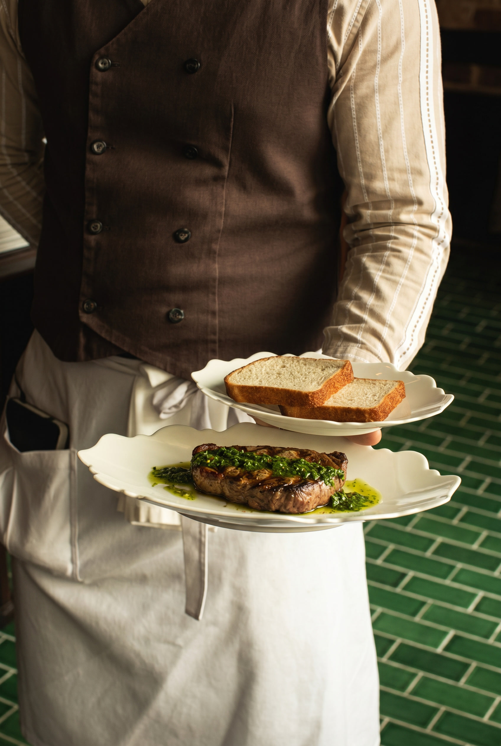 Waiter holding two white plates, one with two slices of bread and the other with a grilled steak topped with green herb sauce.