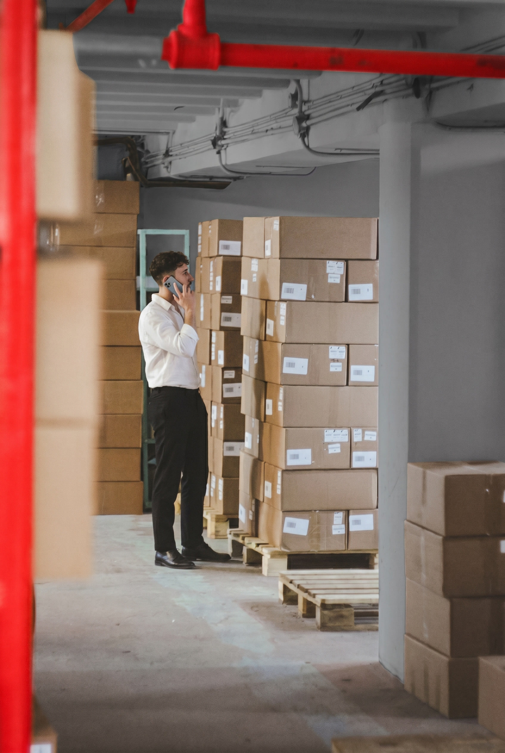 Man in white shirt and black pants talking on phone standing next to stacked cardboard boxes on wooden pallets in a warehouse.