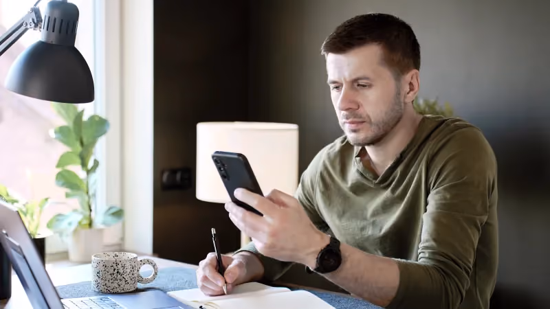 Muž u domácího stolu drží mobil a druhou rukou si píše poznámky do sešitu, vedle notebook a hrnek s kávou.