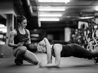 A fitness trainer watches and guides a woman performing a plank exercise in a modern gym. Black-and-white photo with bright overhead lighting and workout equipment in the background.