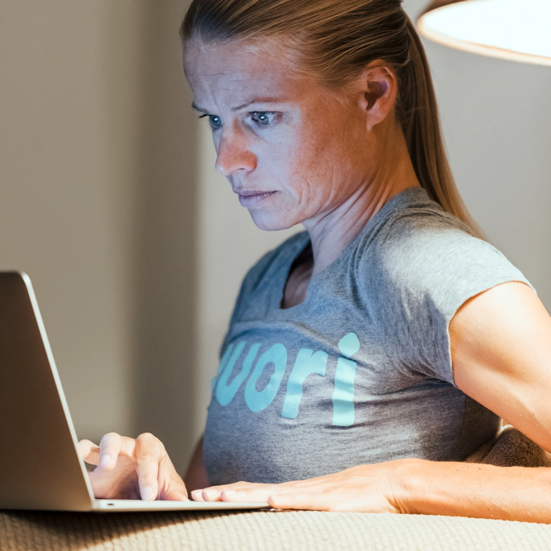 Helle Frederiksen wearing a gray shirt focused on typing on a laptop indoors with warm lighting.