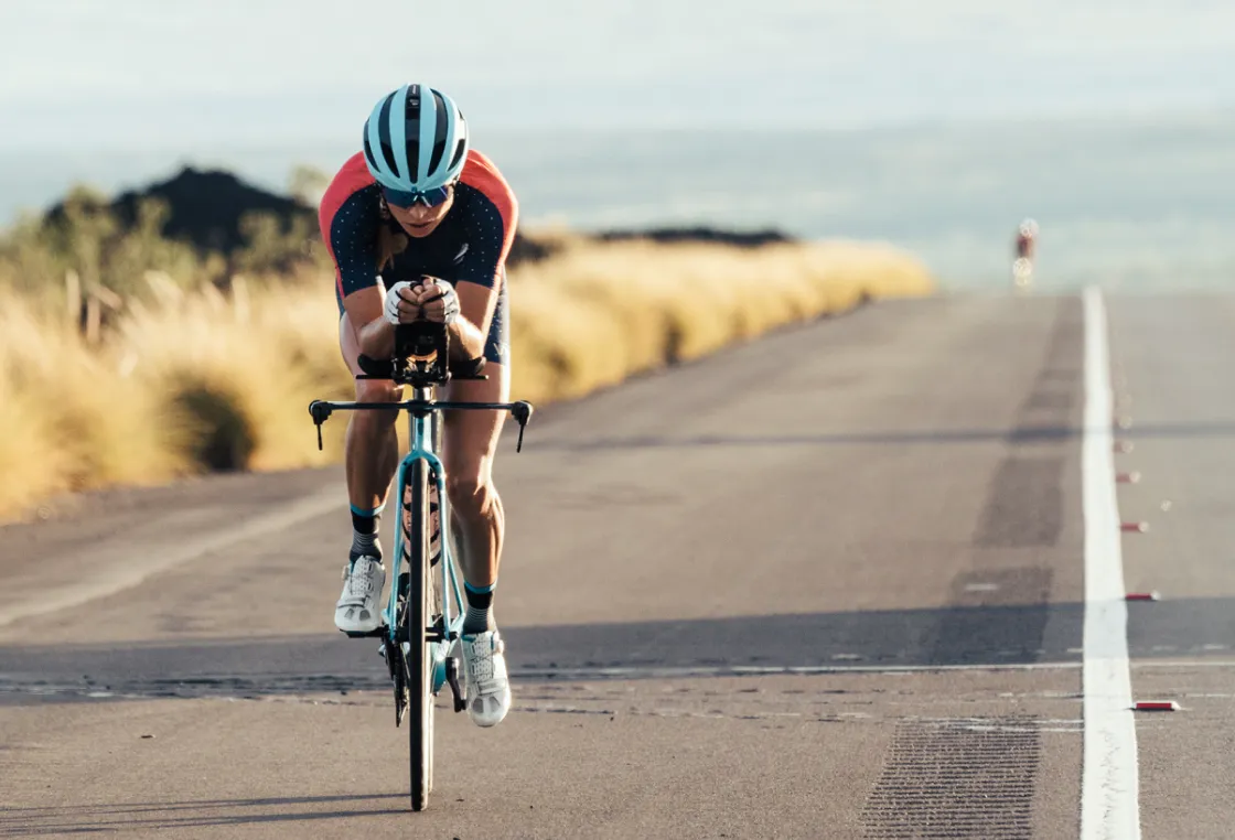 Helle Frederiksen in aerodynamic position wearing a blue helmet and red and blue jersey riding on a road with blurred vegetation in the background.