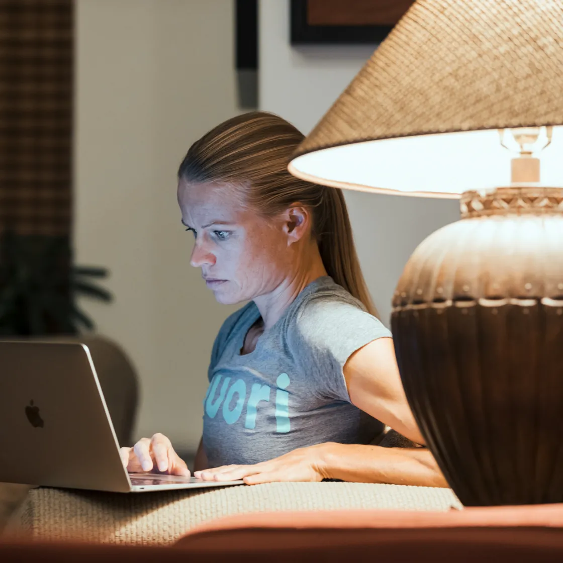 Helle Frederiksen sitting on a couch using a laptop, illuminated by a nearby table lamp.